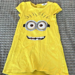 Gul klänning med Minion-tryck  - Säljer en söt gul klänning från H&M med ett stort Minion-tryck på framsidan. Klänningen har korta ärmar och är perfekt för alla Minion-fans. Den är gjord i mjukt bomullsmaterial och har en bekväm passform.