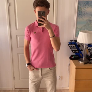 Rosa pikétröja från Lyle & Scott - Säljer en snygg rosa pikétröja från Lyle & Scott med klassisk krage och knappar framtill. Tröjan har en broderad logga på bröstet och är perfekt för en stilren look. Passar både till jeans och chinos! 183cm på bilden!