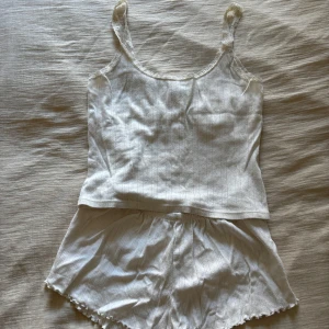 Vit ribbad pyjamas med volangkant - Säljer en söt vit pyjamas i ribbad bomull med diskret hålmönster och volangkant nertill. Shortsen har elastisk midja och är perfekta för varma dagar eller som mysplagg hemma. Matchar toppen med spetsdetaljer om du vill ha ett set.