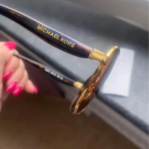 Runda solglasögon från Michael Kors - Snygga runda solglasögon från Michael Kors med tunna guldfärgade metallbågar och bruna tonade glas. Skalmarna är mörkbruna med gulddetaljer och logga på sidan. Perfekta för att lyfta din sommarstil.