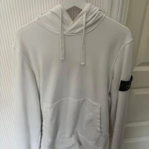 Vit hoodie från Stone Island - Vit hoodie från Stone Island med klassisk patch på ärmen. Tröjan har huva med snörning och en stor magficka framtill. Tillverkad i mjuk bomull, perfekt för en clean och stilren look.