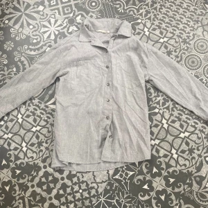 Grå skjorta i bomull med knappar - Stilren grå skjorta i bomull med klassisk krage och knäppning framtill. Skjortan har två bröstfickor och långa ärmar. ☀️