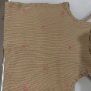Beige ribbad linnetopp med hjärtan - Supersöt beige ribbad linnetopp med tunna axelband och små broderade rosa hjärtan över hela toppen. Perfekt för varma dagar och ger en gullig vibe till din outfit. Toppens material är mjukt och stretchigt för en bekväm passform.