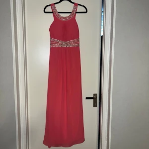 Lång rosa klänning med pärlor - Snygg långklänning i stark rosa med breda axelband och markerad midja. Klänningen har glittriga detaljer med pärlor och strass runt halsringningen och midjan. Tyget är lätt och faller fint, perfekt för bal eller fest. Riktigt statement-plagg för dig som vill sticka ut. Lite pärlor som hänger men det går att fixa ganska enkelt💗