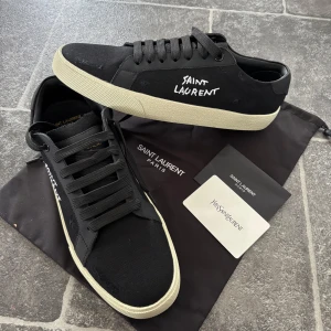 Yves Saint Laurent skor  - Svarta sneakers från Saint Laurent med vit sula och broderad logga på sidan. Skorna har rund tå, platt sula och snörning. Kommer tyvärr utan box då jag har tappat bort det. För mer bilder tveka inte skriva. Och med snabb äffar så löser vi något fint.. Snygg minimalistisk design som passar till allt.