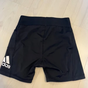 Svarta träningstights-shorts från Adidas - Svarta träningstights-shorts från Adidas med vit logga på benet. De har hög midja, elastiskt band och en praktisk ficka på sidan. Snygg och sportig passform i stretchigt syntetmaterial, perfekta för gymmet eller löprundan.