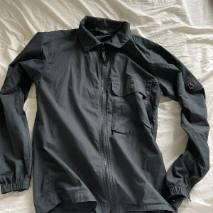 Svart overshirt - Svart overshirt från Lagency med bröstficka med knapp och patch på ärmen. Jackan har klassisk krage och perfekt för lager-på-lager och enkel att styla till olika outfits. Ny skick, storlek small