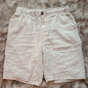 Beige linneshorts från H&M med snörning. Storlek 158 - Snygga beige shorts från H&M i en skön mix av linne och bomull. De har elastisk midja med både snörning och knapp, samt fickor fram och en bakficka. Perfekta för varma dagar och chill stil. Lätta att matcha med allt.