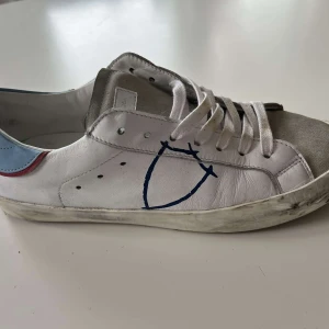 Philippe Model vita sneakers med blå detalj - Snygga vita Philippe Model sneakers med blå och röda detaljer på hälen och grå mocka framtill. Skorna har snörning, platt sula och en handritad blå logga på sidan. Tillverkade i skinn och mocka för en cool och urban look.