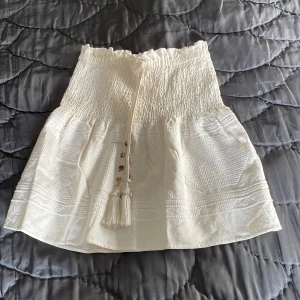  Zarakjol - Supersöt vit kortkjol från Zara med smockad midja, volangkant upptill och dekorativa tofsar med pärlor framtill. Kjolen har ett lätt strukturerat tyg och känns perfekt för varma dagar. Passar dig som gillar en feminin och trendig stil.