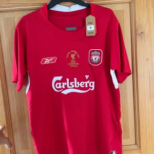 Liverpool Gerrard 8 Champions League tröja - Liverpool FC matchtröja från Champions League-finalen 2005 med Gerrard 8 på ryggen. Röd med vita detaljer, broderade klubb- och turneringsmärken samt Carlsberg-tryck på bröstet. Tillverkad i ventilerande polyester, officiell team merchandise.