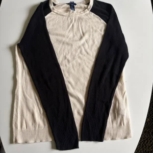 Beige och svart stickad tröja Chaps - Snygg stickad tröja från Chaps i beige med svarta ärmar. Tröjan har rund halsringning, ribbade muddar och långärmad design. Perfekt för dig som gillar en stilren och enkel look med kontrastfärger. Storlek L men passar M