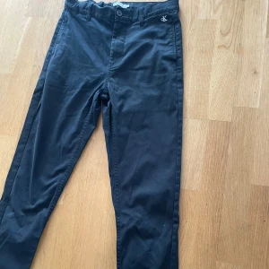 Svarta chinos från Calvin Klein - Svarta chinos från Calvin Klein med diskret CK-brodyr framtill och logga bak. Klassisk rak passform med bälteshällor och knappgylf. Snygga bakfickor och mjukt bomullsmaterial som känns skönt hela dagen.
