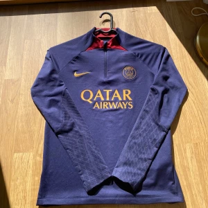 PSG långärmad träningströja Nike 147-158 - Snygg långärmad PSG träningströja från Nike i mörkblått med gul logga och tryck. Tröjan har halv dragkedja, röd detalj i nacken och är gjord i Dri-FIT material som andas. Perfekt för fotbollsträning eller för vanlig träning.
