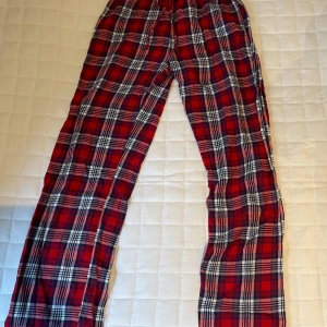 Rödrutiga mjukisbyxor med snörning - Snygga och sköna mjukisbyxor i röd, svart och vit rutig design. Byxorna har elastisk midja med snörning för extra komfort. Perfekta för chill dagar hemma eller när du vill vara bekväm men ändå se cool ut. Lappen är bort klippt men dom är i storlek 170 från ginas barnavdelning 