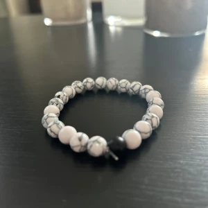 Vitt pärlarmband med marmormönster - Stilrent armband med runda vita pärlor och grått marmormönster, samt en svart pärla som accent. Elastiskt band gör det enkelt att ta på och av. Perfekt accessoar för att lyfta din outfit med en cool och modern vibe.