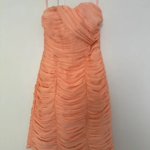 Persikofärgad axelbandlös klänning med drapering - Superfin kort axelbandlös klänning i persikofärg och snygg omlott-detalj upptill. Klänningen har draperad design över hela och är figurnära med en lätt volym. Perfekt för dig som vill sticka ut med en unik look