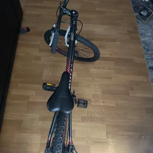 Sports cykel alla åldrar - Bilderna visar en svart och röd mountainbike med grova däck och framdämpare. Cykeln ser robust ut och passar för äventyr och fritidsaktiviteter. Perfekt för dig som gillar att utforska naturen eller cykla i stan. Snygg design som sticker ut och ger en sportig känsla.