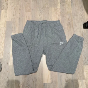 Grå mjukisbyxor från Nike M - Gråa mjukisbyxor från Nike med broderad vit logga på benet. Byxorna har resår och snörning i midjan samt muddar vid bensluten. Tillverkade i mjukt bomullsmaterial för maximal komfort. Perfekta för chill eller träning.