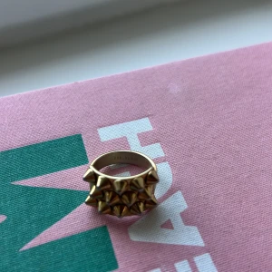 Guldig statementring med nitar - Cool och chunky ring i guldton med flera rader av spetsiga nitar framtill. Ringen har en bred design som verkligen sticker ut och ger edgy vibes. Perfekt accessoar för dig som vill ha något unikt och vågat.