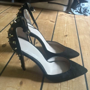 Svarta pumps med nitar och spetsig tå - Svarta klackar i mocka med spetsig tå och hög klack. coola detaljer med guldnitar och öglor på hälen och klacken. Rem runt vristen med spänne. 