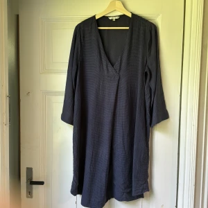 Mörkblå klänning från Indiska L - En stilren mörkblå klänning från Indiska, modell Serenity. Klänningen har v-ringning, trekvartsärm och är tillverkad i ett lätt, randigt tyg. Passformen är loose och längden är något över knäet. Perfekt för dig som gillar enkel och avslappnad stil.