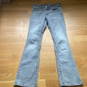 Grå bootcut jeans från H&M - Snygga grå jeans från H&M med bootcut-ben och låg midja. Jeansen har justerbar midja, klassiska fem fickor och coola detaljer med knappar och bälteshällor. Perfekta för dig som gillar en avslappnad men trendig look.