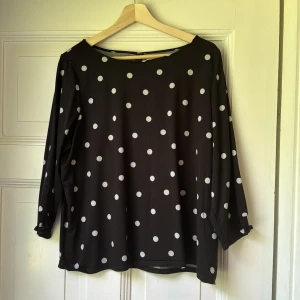 Svart prickig blus med trekvartsärm- Bondelid - Svart blus med vita prickar och trekvartsärmar. Blusen har en rak passform, rund halsringning och diskret resår vid ärmsluten. Materialet känns lätt och flowy, perfekt för dig som gillar en stilren men lekfull look. Märke Bondelid