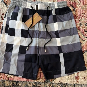 Burberry rutiga badshorts med snörning - Snygga badshorts från Burberry med klassiskt rutigt mönster i svart, grått och vitt. Shortsen har elastisk midja med svart snörning och metalländar med Burberry-logga. Perfekta för sommaren och strandhäng, med praktiska fickor på sidorna.