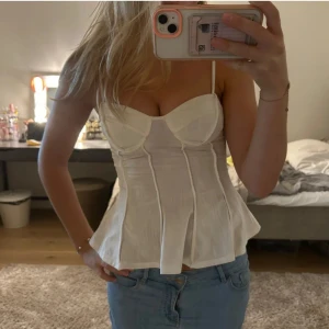 Vit korsett-topp med tunna axelband - Supersnygg vit korsett-topp med tunna axelband och markerade sömmar som ger en riktigt trendig siluett. Toppen har en lätt utsvängd nederdel och en figurnära passform som framhäver midjan. Perfekt för dig som vill ha en feminin och modern look.