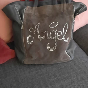 Stor brun shoppingväska med text - Säljer en stor och rymlig shoppingväska i brunt och grått med texten 'Angel' i glittriga nitar på framsidan. Väskan har två handtag och dragkedjor på sidorna. Perfekt för dig som gillar statement-accessoarer.