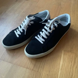 Svarta Saint Laurent - Svarta sneakers från Saint Laurent i mocka med vita skinnpaneler på hälen och vita snören. Klassisk låg modell med rund tå och platt vit sula. Snygg och stilren design med diskret logga på baksidan och sulan.