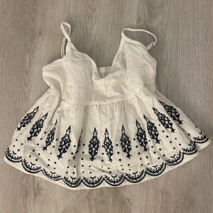 Vit broderad linnetopp med spetskant - Supersöt vit linnetopp med tunna axelband och v-ringning. Toppen har mörkblå broderade detaljer och prickar längs den vågiga spetskanten nertill. Perfekt för varma dagar och ger en bohemisk vibe.