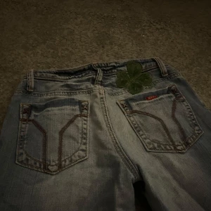 Miss Sixty ljusblå bootcut jeans - Säljer ett par ljusblå bootcut jeans från Miss Sixty med låg midja. Jeansen har klassiska bakfickor med kontrastsömmar och en grön fyrklöver-detalj ovanför ena fickan. Materialet är denim och passformen är loose med utsvängda ben. Uppklippta pg av slitningar 😗