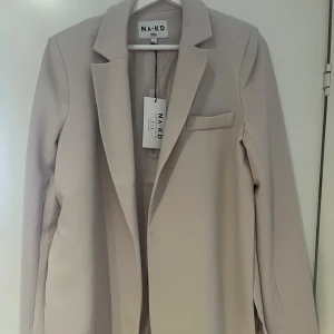 Beige kavaj från NA-KD, stl 36 - Stilren beige kavaj från NA-KD i storlek 36. Klassisk modell med krage, långa ärmar och en bröstficka. Snyggt fodrad och i ett mjukt, slätt material som ger en clean look. Perfekt att styla med jeans eller kostymbyxor. Har slits på sidorna. Storlek 36 men jag har vanligt vis 40/42 och den passar mig. Beror på passform. 