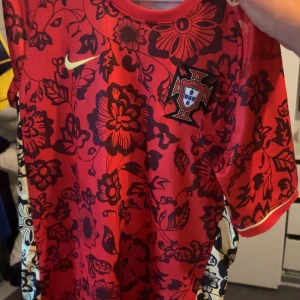 Portugal Nike röd blommig fotbollströja - Snygg röd Portugal fotbollströja från Nike med svart blommigt mönster över hela tröjan. Tröjan har korta ärmar och det portugisiska landslagets emblem på bröstet. Perfekt för dig som vill sticka ut på match eller i vardagen.