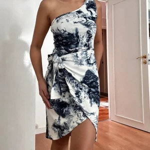 Oneshoulder kortklänning med knyt - Snygg kortklänning med oneshoulder och coolt blåvitt mönster. Klänningen har omlottkänsla med knyt i midjan och en asymmetrisk nederkant. Perfekt för dig som vill sticka ut med en unik look.