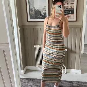 Randig ribbad långklänning - Säljer en färgglad ribbad långklänning med smala axelband och öppen rygg. Klänningen har ett mönster av smala ränder i grönt, blått, orange, vitt och gult. Den är figurnära och har en mjuk, stretchig känsla som passar perfekt till sommaren.