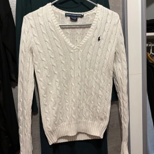 Vit kabelstickad tröja Ralph Lauren - Vit kabelstickad tröja från Ralph Lauren med v-ringning och broderad logga på bröstet. Tröjan har lång ärm och ribbade muddar, perfekt för lager-på-lager och en klassisk preppy stil.