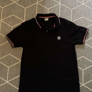 Svart Moncler pikétröja  - Svart pikétröja från Moncler med klassisk krage och korta ärmar. Tröjan har Moncler-logga på bröstet och snygga rödvita detaljer på krage och ärmslut. Tillverkad i 100% bomull för en skön och stilren känsla.