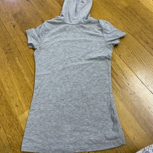 Grå topp  med huva Terranova - Superenkel och chill grå topp från Terranova i mjuk bomull. Toppen har korta ärmar och en huva som ger en sportig vibe. Perfekt för dig som gillar en avslappnad och stilren look.