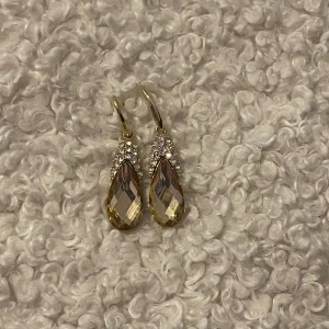 Guld örhängen med droppformad kristall - Snygga örhängen med droppformade, facetterade kristaller i guldton. Ovanför kristallen sitter små gnistrande stenar som ger extra bling. Krokar i guldfärg. Perfekta för dig som vill sticka ut med lite extra glans.
