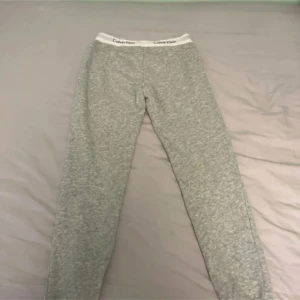 Grå mjukisbyxor från Calvin Klein - Snygga grå mjukisbyxor från Calvin Klein med vit resår i midjan där loggan syns tydligt. Byxorna har raka ben och är gjorda i mjukt bomullsmaterial, perfekta för chill dagar. Klassisk och enkel design som funkar till allt.