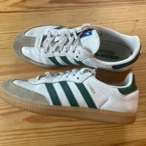 Adidas Samba vita och gröna sneakers - Klassiska Adidas Samba sneakers i vitt skinn med gröna ränder och detaljer. Skorna har beige mockadetaljer vid tån och en brun gummisula med mönster. Ikonisk Samba-logga i guld på sidan och snörning framtill. Perfekta för en sportig och retro look.
