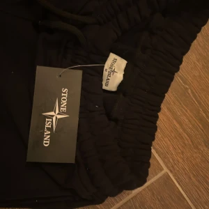 Svarta shorts från Stone Island som kommer med pris lapp - Svarta shorts från Stone Island med klassisk logga på benet. Modellen har elastisk midja med snörning och praktiska fickor på sidorna. Sköna och stilrena shorts i mjukt bomullsmaterial, perfekta för en avslappnad look. Plagget kommer med prislappen