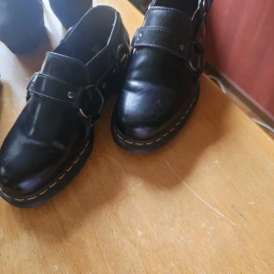 Svarta Dr. Martens loafers i skinn - Klassiska svarta loafers från Dr. Martens i blankt skinn med bred rem och metallspännen. Snygga kontrastsömmar runt sulan och robust, räfflad yttersula. Perfekta för dig som gillar en edgy och tidlös stil.