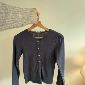 Mörkblå kofta med spetsdetalj - Supersöt mörkblå kofta från NoaNoa med lång ärm och fina spetsdetaljer vid halsringningen. Koftan har dekorativa knappar i olika färger och ett diskret hålmönster i tyget. 