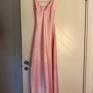 Lång rosa satinklänning med smala band - Supersnygg lång klänning i ljusrosa satin med smala axelband och v-ringning fram. Klänningen har öppen rygg med snygga banddetaljer och ett elegant!🩷 Nypris:399kr