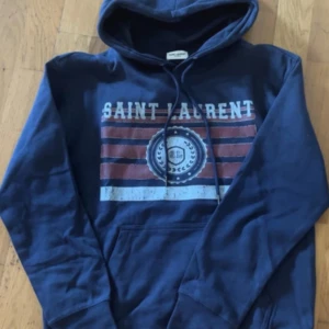 Saint Laurent  - Säljer en blå hoodie från Saint Laurent med tryck framtill i vitt och rött. Tröjan har huva med snörning och en stor magficka. Perfekt för dig som gillar streetstyle och vill ha något bekvämt och snyggt. Storlek är M men passar på S runt 174 cm