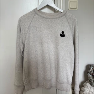 Grå sweatshirt från Isabel Marant - Snygg ljusgrå sweatshirt från Isabel Marant med svart logga på bröstet. Tröjan har rund halsringning, långa ärmar och är tillverkad i mjuk bomull. Perfekt för en chill och stilren look. Ny skick 